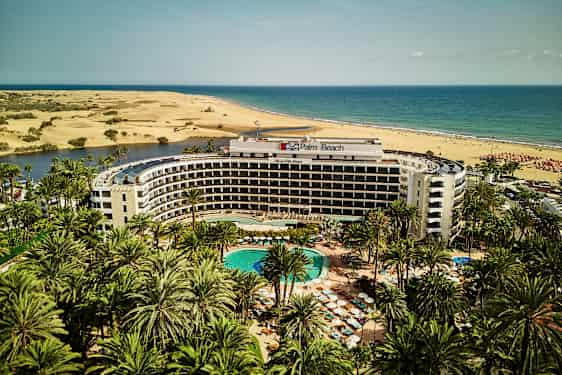 Palm Beach hotell med basseng i forgrunnen, omgitt av palmer, med sanddyner og hav i bakgrunnen. (AI-generert tekst)