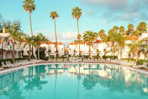 Hotellområde med hvitkalkede bygninger med røde tak, omgitt av palmer, og et stort basseng med solsenger rundt. (AI-generert tekst)