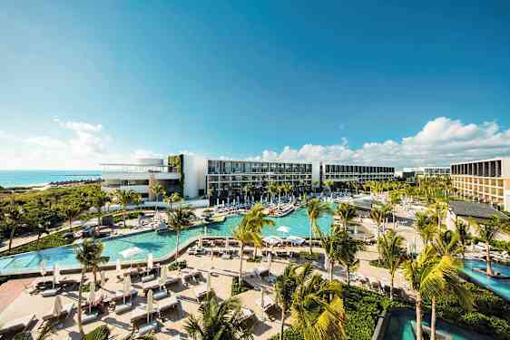 Et moderne hotellkompleks med flere bassenger, frodig vegetasjon og palmer, beliggende ved kysten med utsikt over havet under en klar blå himmel. (AI-generert tekst)