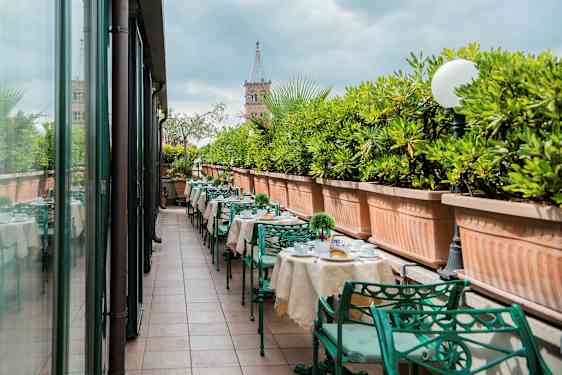 En utendørsterrasse med dekkede bord og grønne metallstoler, omgitt av store plantekasser med grønne planter. (AI-generert tekst)