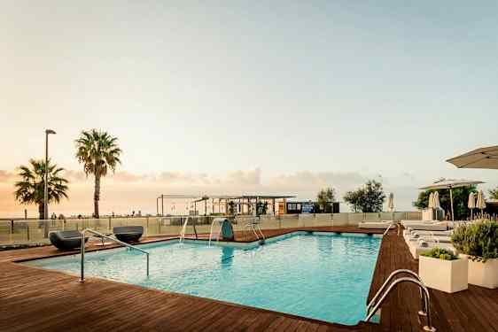 Bildet viser et utendørs svømmebasseng med treterrasse og solsenger ved kanten. I bakgrunnen er noen palmer, en bygning og havet under en lys kveldshimmel. (AI-generert tekst)