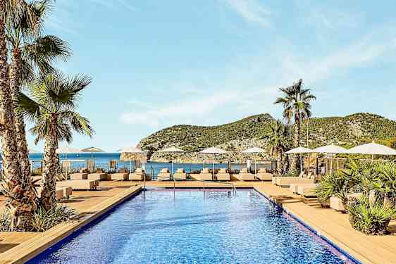 Et basseng med solsenger og parasoller, omgitt av palmer, med utsikt over havet og fjellene i bakgrunnen. (AI-generert tekst)