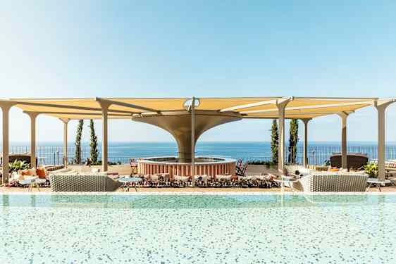 Et basseng med utsikt over havet og møblerte områder under skyggefulle tak. (AI-generert tekst)