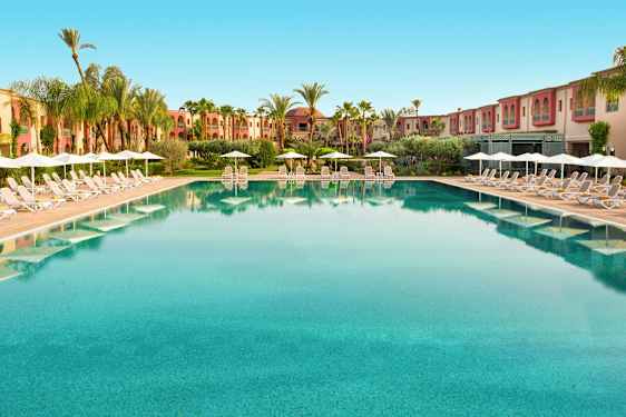 Stor basseng med solsenger og parasoller langs kantene, omgitt av palmer og bygninger. (AI-generert tekst)