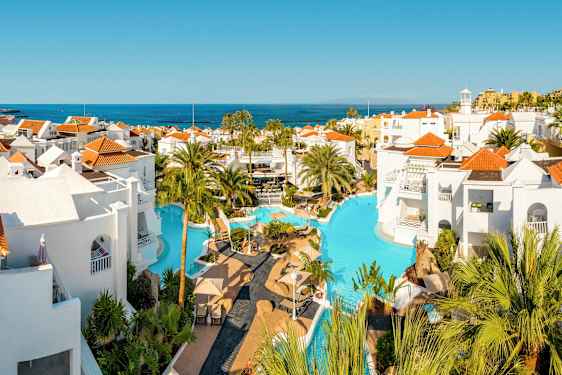 Bassengområde med hvite bygninger og røde tak, omgitt av palmer med utsikt over havet. (AI-generert tekst)