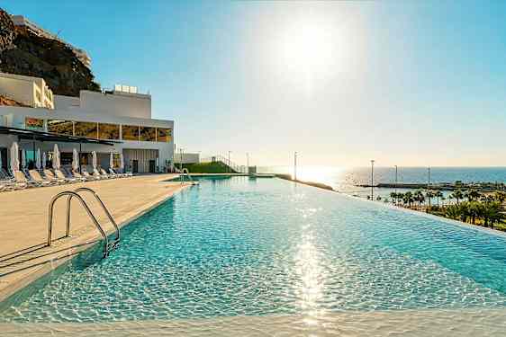 Et infinity-basseng med speilklart vann, solsenger og parasoller på siden, i et hotellområde med utsikt mot havet og stranden. (AI-generert tekst)