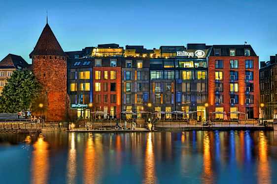 En historisk murbygning ved en moderne hotell ved vannet om kvelden. (AI-generert tekst)