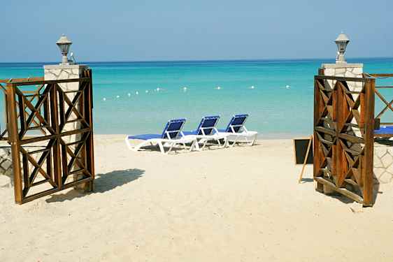 En sandstrand med solsenger og en åpen port med utsikt over det turkise havet. (AI-generert tekst)