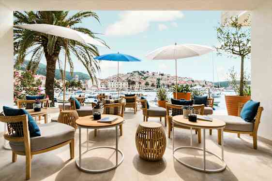 Utendørs serveringsområde med bord og stoler under parasoller, omgitt av palmer, med utsikt over en pittoresk havn og by. (AI-generert tekst)