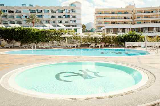 To svømmebassenger omgitt av solsenger og parasoller, med hotellbygninger i bakgrunnen. (AI-generert tekst)