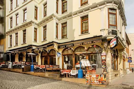 En gate med en bygning som har restaurant med uteservering, skilt med mat som kebab og pizza. (AI-generert tekst)