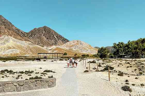 Et tørt landskap med personer som går på en sti som fører mot noen små bygninger. Fjellet er synlig i bakgrunnen under en klar blå himmel. (AI-generert tekst)