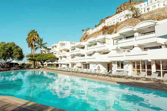 Et basseng foran et hvitt hotellkompleks med flere balkonger. I bakgrunnen vises palmer og en skråning. (AI-generert tekst)