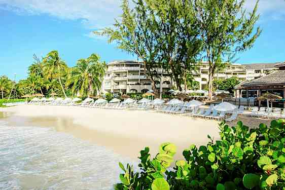 En strand med solsenger og parasoller foran en resort med palmer og beplantning. (AI-generert tekst)