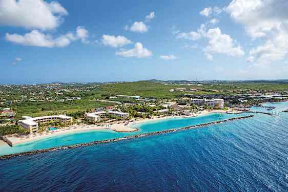 Luftfoto av en kystlinje med hotellbygninger ved en strand og grøntområder i bakgrunnen. (AI-generert tekst)