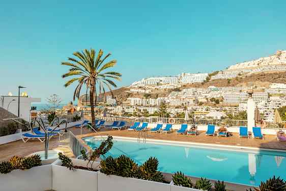 Utendørsbasseng med solsenger og palmer samt utsikt over fjell og bygninger. (AI-generert tekst)