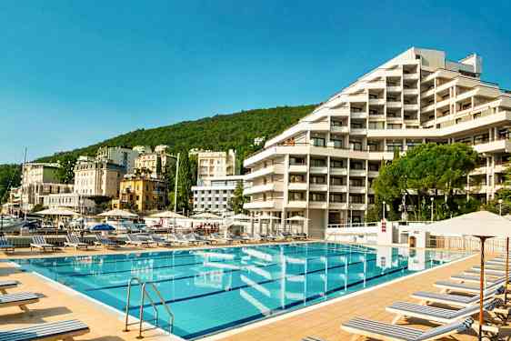 Bassengområde med solsenger foran et hotell, omgitt av grønne åser og flere bygninger i bakgrunnen. (AI-generert tekst)