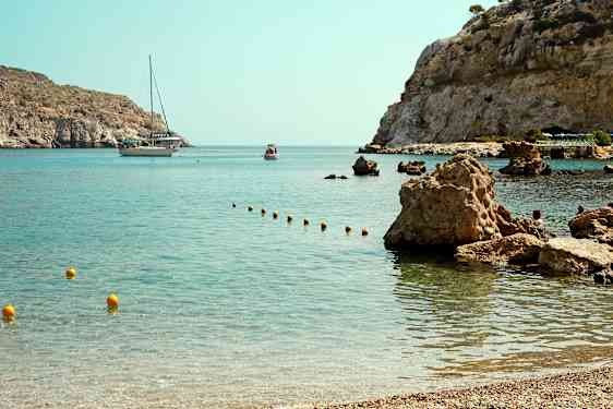 En bukt med klart vann og steinete kyst, hvor en båt og en mindre flytende fartøy er synlige i bakgrunnen. (AI-generert tekst)