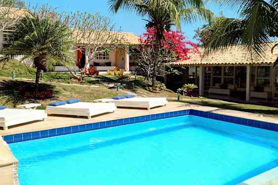 Et basseng omgitt av solsenger og tropiske planter ved en bygning med terrakottatak. (AI-generert tekst)