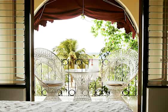 Utsikt fra et rom mot en terrasse med to hvite stoler og et bord med glass, omgitt av grønt og palmer. (AI-generert tekst)