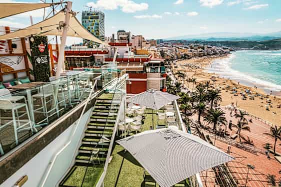 En takterrasse med bord og parasoller, omgitt av glasrekkverk, med utsikt over en strand og byen ved havet. (AI-generert tekst)