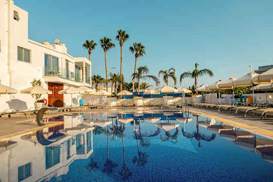 Bilde av et utendørs basseng omgitt av solsenger og parasoller, med palmer og en hvit bygning i bakgrunnen. (AI-generert tekst)