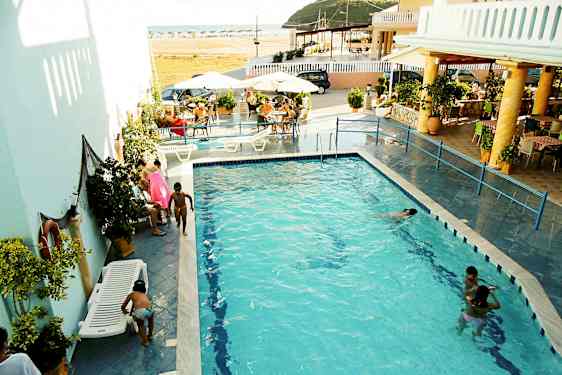 Et utendørs basseng omgitt av solsenger og bord, med folk som svømmer og sosialiserer. (AI-generert tekst)