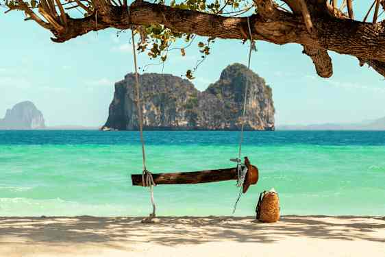 En trehuske henger fra et tre ved en sandstrand med havet og klippeformasjoner i bakgrunnen. (AI-generert tekst)