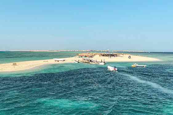 En liten sandøy med båter ved brygga, omgitt av klart blått vann. (AI-generert tekst)