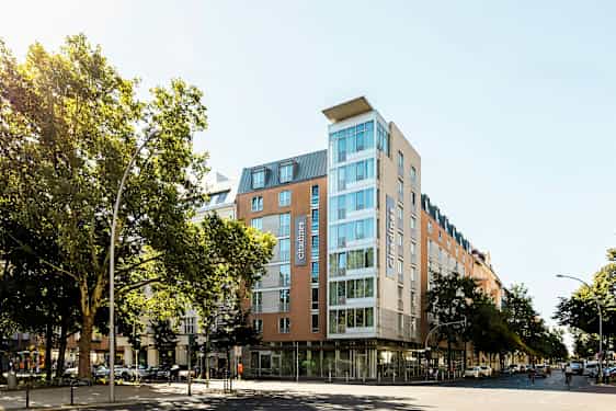 Et moderne hotell med glassfasade omgitt av trær og bymiljø. (AI-generert tekst)