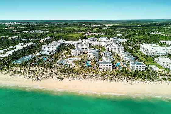 Flyfoto av et hotellkompleks ved en strand som inkluderer bassengområde, omgitt av grønt. (AI-generert tekst)