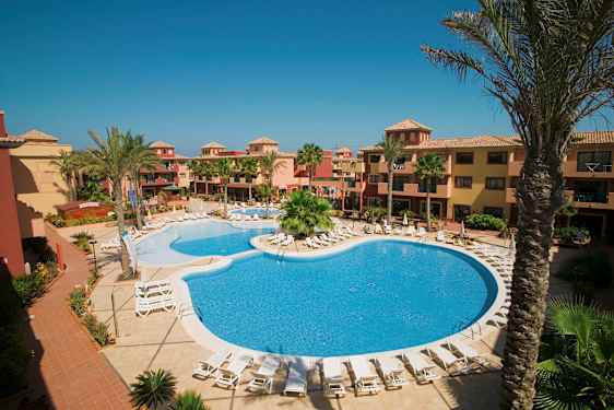 Hotellområde med basseng og solstoler, omgitt av palmer og bygninger i varme farger. (AI-generert tekst)