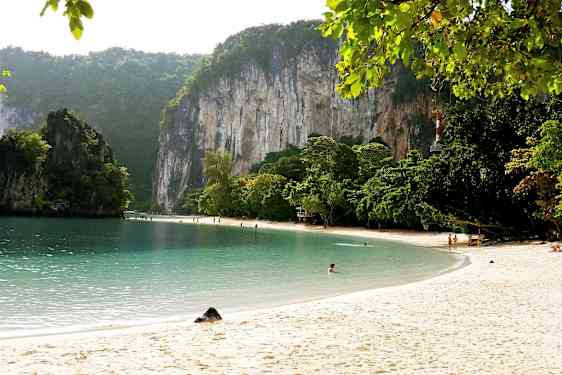 Sandstrand med klart vann, omgitt av grønne trær og høye kalksteinsklipper. (AI-generert tekst)