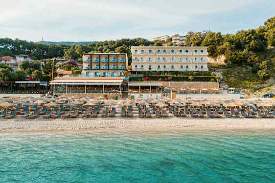 En sandstrand ved havet med solsenger og parasoller. Bakgrunnen viser hotellbygninger og vegetasjon. (AI-generert tekst)