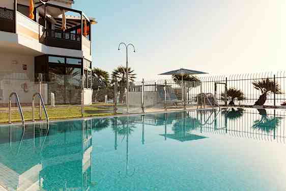 Et basseng omgitt av et glassgjerde med utsikt mot en plen og solsenger under parasoller. (AI-generert tekst)