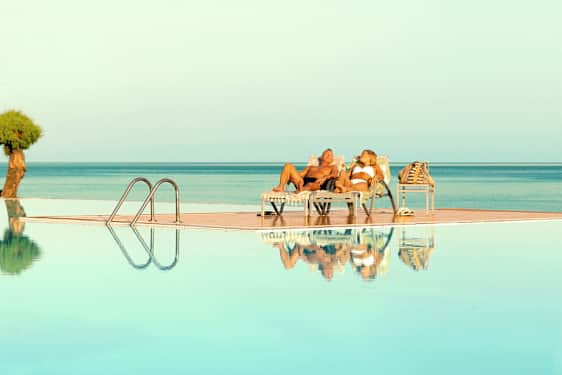 To personer ligger på solsenger ved et basseng med utsikt over havet. (AI-generert tekst)