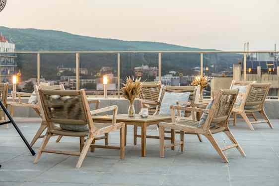 Terrasse med en gruppe tremøbler, inkludert stoler og bord. Utsikt over byen og grønne åser i bakgrunnen. (AI-generert tekst)