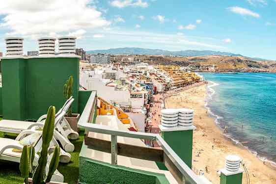 Utsikt fra en takterrasse med solsenger mot en strand og bymiljø. (AI-generert tekst)