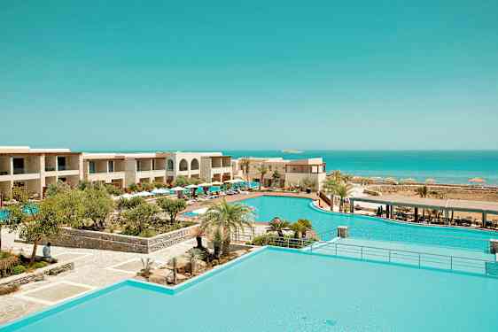 Et ferieanlegg med bassengområde omgitt av tropiske planter og bygninger, med utsikt over havet. (AI-generert tekst)