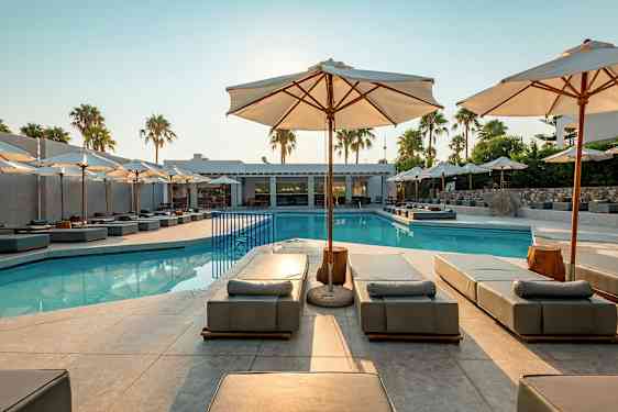Utendørsbasseng med solsenger og parasoller, omgitt av palmer og bygninger. (AI-generert tekst)