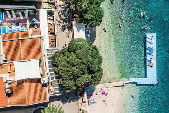 Flyfoto av bygninger med røde tak, et basseng, en strand og turkist hav. (AI-generert tekst)