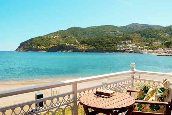 Utsikt fra en balkong mot havet og frodige åser, med en trestol og et bord. (AI-generert tekst)