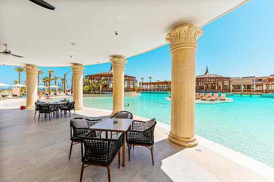 Utendørs serveringsområde med bord og stoler ved et basseng, omgitt av søyler og solsenger. (AI-generert tekst)
