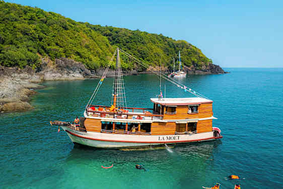 En trebåt ved navn La Moet ligger fortøyd i turkist vann nær en skogkledd kyst. (AI-generert tekst)