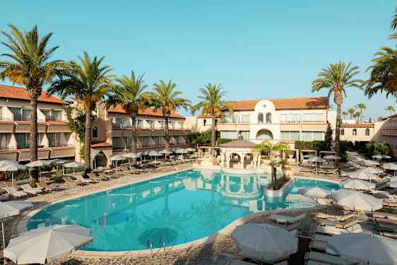 Hotellbygning med solsenger og parasoller rundt et utendørsbasseng, omgitt av palmer. (AI-generert tekst)