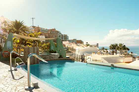 Et infinity-basseng med utsikt over hustak, palmer og havet i et solrikt landskap. (AI-generert tekst)