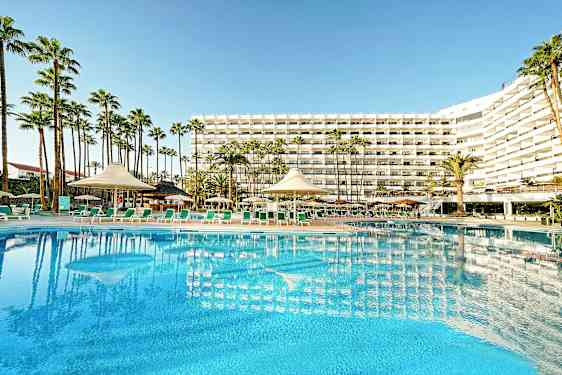 Hotellbygning med stort basseng og solsenger i forgrunnen, omgitt av palmer. (AI-generert tekst)