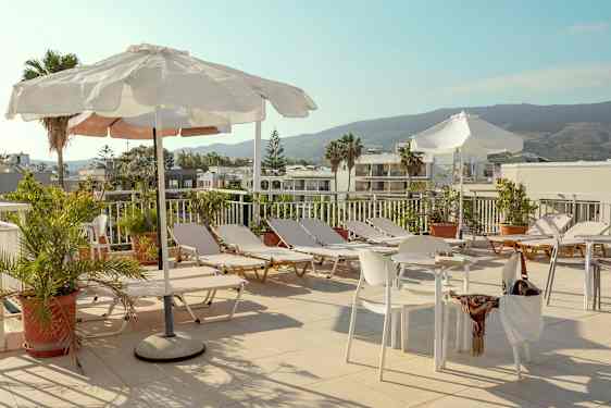 En solterrasse med solsenger, parasoller og bord samt stoler omgitt av grøntområder med utsikt mot bygninger og fjell. (AI-generert tekst)