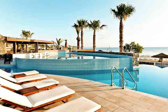 Et basseng med solsenger i kanten, palmer og havet i bakgrunnen. (AI-generert tekst)