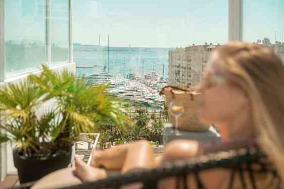 Utsikt mot båter i en marina, med en glassbalkong og frodige planter i forgrunnen. (AI-generert tekst)